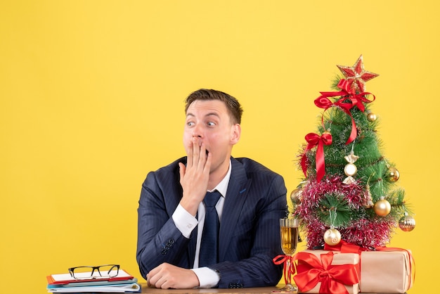 Vista frontal asombrado hombre sentado en la mesa cerca del árbol de navidad y presenta sobre fondo amarillo espacio libre