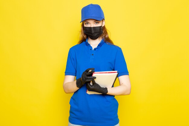 Una vista frontal atractiva mujer mensajero en polo azul gorra azul y jeans en guantes negros máscara protectora negra con pluma y cuaderno sobre el fondo amarillo trabajo de servicio de alimentos