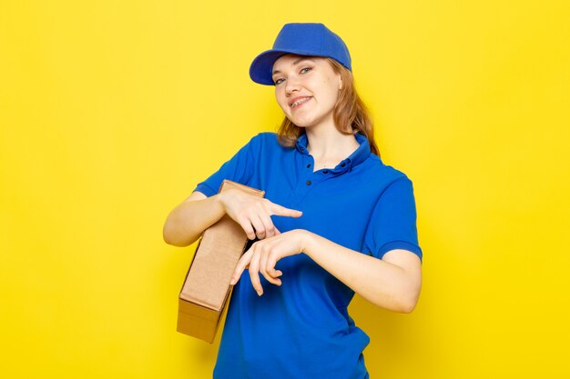 Una vista frontal atractiva mujer mensajero en polo azul gorra azul y jeans sosteniendo paquete tocando su muñeca sonriendo en el trabajo de servicio de alimentos de fondo amarillo