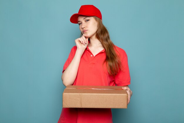 Una vista frontal atractiva mujer mensajero en polo rojo gorra roja y jeans sosteniendo paquete posando pensando en el trabajo de servicio de alimentos de fondo azul