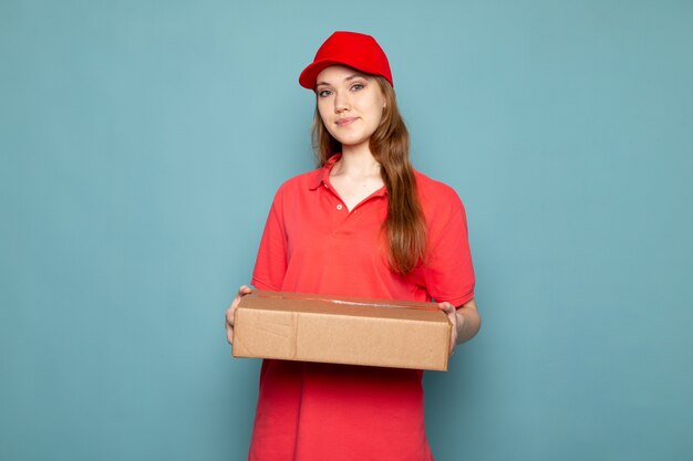 Una vista frontal atractiva mujer mensajero en polo rojo gorra roja y jeans sosteniendo paquete posando sonriendo sobre el fondo azul trabajo de servicio de alimentos