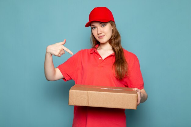 Una vista frontal atractiva mujer mensajero en polo rojo gorra roja y jeans sosteniendo paquete posando sonriendo sobre el fondo azul trabajo de servicio de alimentos