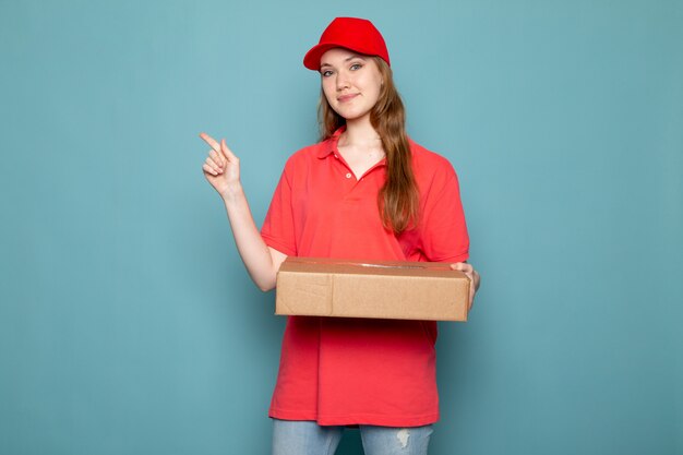 Una vista frontal atractiva mujer mensajero en polo rojo gorra roja y jeans sosteniendo paquete posando sonriendo sobre el fondo azul trabajo de servicio de alimentos