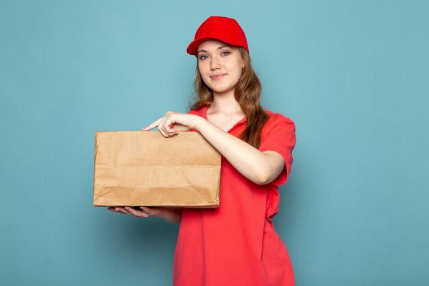 Una vista frontal atractiva mujer mensajero en polo rojo gorra roja y jeans sosteniendo paquete posando sonriendo sobre el fondo azul trabajo de servicio de alimentos