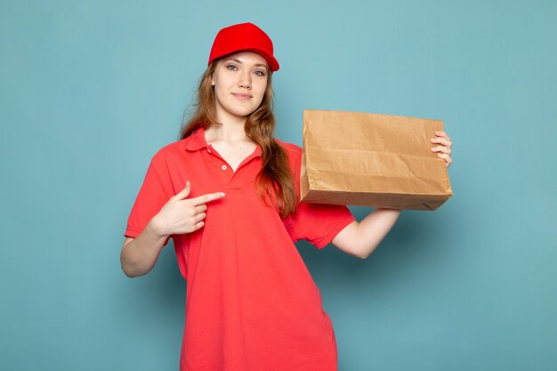 Una vista frontal atractiva mujer mensajero en polo rojo gorra roja y jeans sosteniendo paquete posando sonriendo sobre el fondo azul trabajo de servicio de alimentos