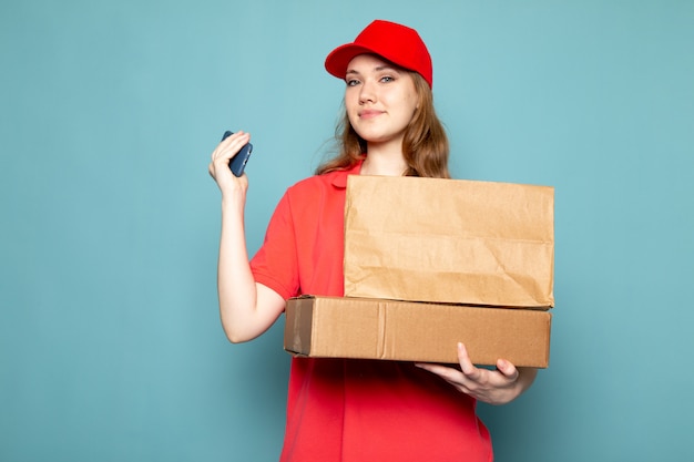 Una vista frontal atractiva mujer mensajero en polo rojo gorra roja con paquete marrón usando su teléfono sonriendo sobre el fondo azul trabajo de servicio de alimentos