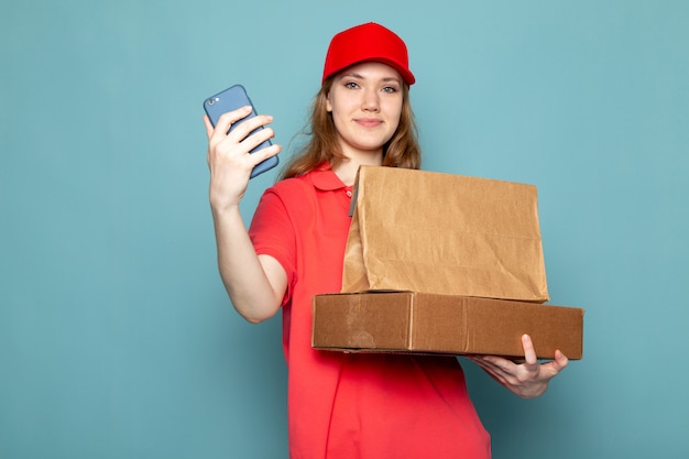 Una vista frontal atractiva mujer mensajero en polo rojo gorra roja con paquete marrón usando su teléfono sonriendo sobre el fondo azul trabajo de servicio de alimentos