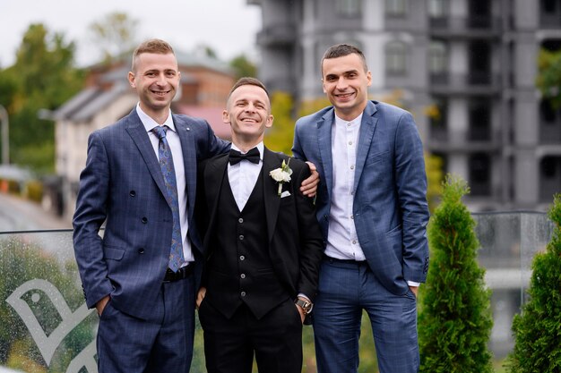 Vista frontal de un atractivo y feliz grupo de amigos con el novio vestido con ropa de boda de moda parados en el balcón al fondo de los edificios y mirando la cámara durante la celebración de la boda