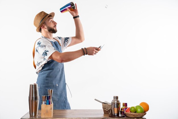 Vista frontal del barman masculino frente a la mesa de la barra preparando una bebida en la coctelera en la pared blanca bar trabajo de alcohol club de frutas bebida nocturna