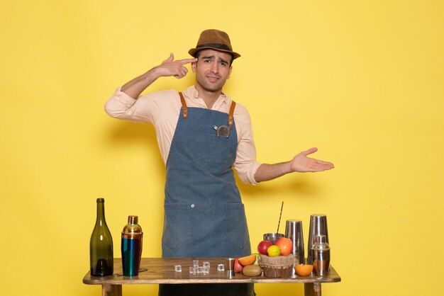 Vista frontal del barman masculino frente a la mesa con bebidas y agitadores en la pared amarilla beber alcohol nocturno bar club de color masculino