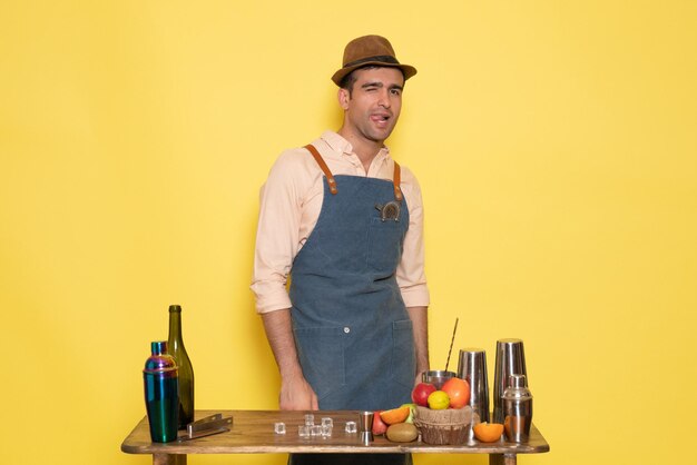 Vista frontal del barman masculino de pie delante del escritorio con bebidas y frutas en la pared amarilla night club bar bebida masculina