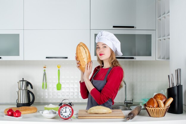 Vista frontal bastante joven en sombrero de cocinero y delantal sosteniendo pan en la cocina