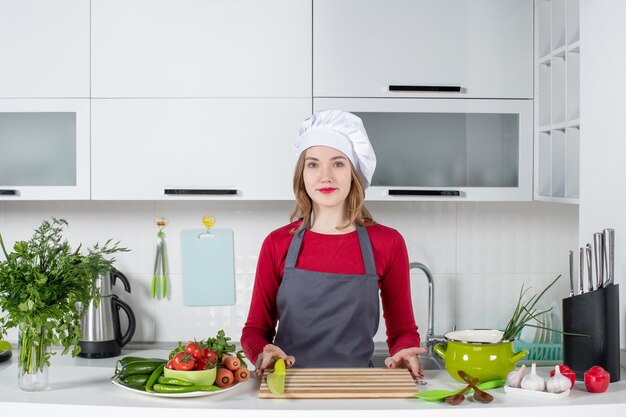 Vista frontal bastante mujer chef en uniforme de pie detrás de la mesa de la cocina