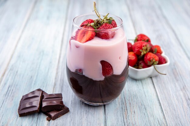 Vista frontal de batido con fresas y chocolate en un vaso con barra de chocolate sobre superficie de madera gris