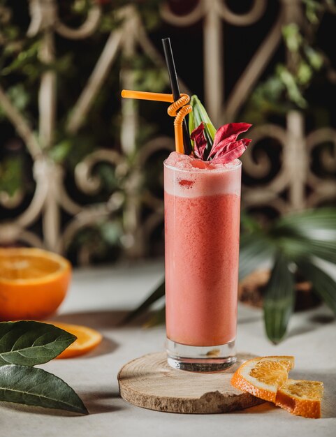 Vista frontal de batido de pomelo rojo en un vaso con paja y rodajas de naranjas sobre un soporte de madera sobre la mesa