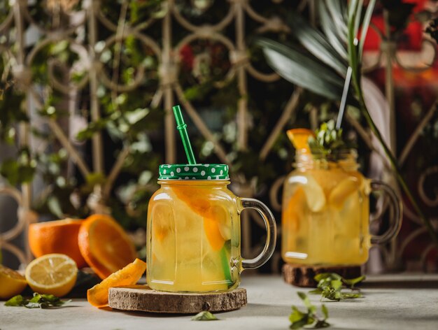 Vista frontal de la bebida de limonada con rodajas de naranja y limón en copa de cóctel con mango y paja sobre un soporte de madera sobre la mesa