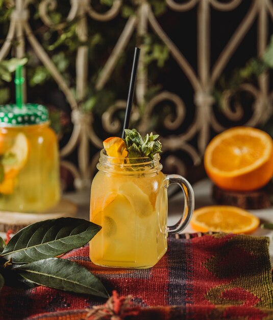 Vista frontal de la bebida de limonada con rodajas de naranja y limón decorado con menta en copa de cóctel con mango y paja sobre un mantel