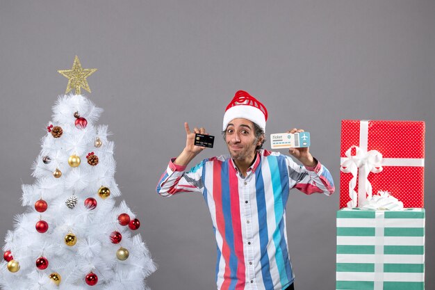 Vista frontal bendito joven sosteniendo la tarjeta y el boleto de viaje alrededor del árbol de Navidad y regalos