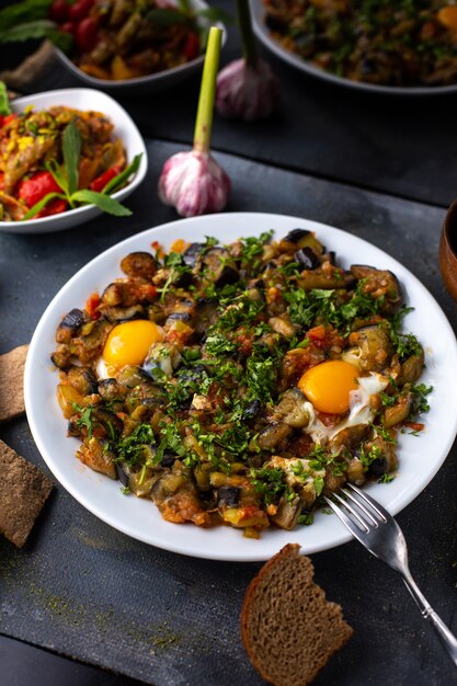 Una vista frontal berenjenas negras mezcladas junto con verduras en rodajas hojas verdes dentro de un plato blanco junto con patatas fritas en el escritorio gris vitaminas vegetales