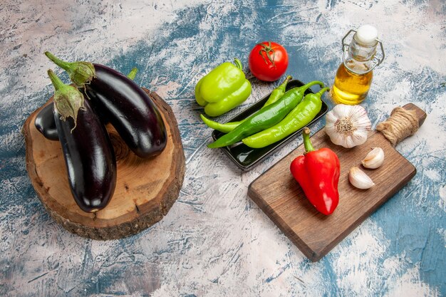 Vista frontal de berenjenas en tablero de madera de árbol pimientos picantes en placa negra tomate pimiento rojo ajo en tabla de cortar berenjena sobre fondo azul-blanco