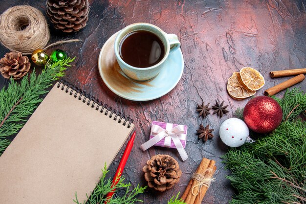 Vista frontal bolígrafo rojo un bloc de notas ramas de pino árbol de navidad juguetes de bolas y taza de regalo de té canela anís hilo de paja sobre fondo rojo oscuro