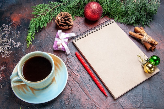 Vista frontal bolígrafo rojo un cuaderno ramas de pino árbol de navidad juguetes de bolas palitos de canela una taza de té sobre fondo rojo oscuro navidad