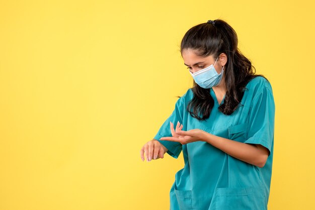 Vista frontal de la bonita doctora con máscara médica comprobando el tiempo en la pared amarilla