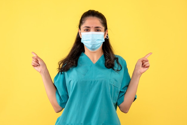 Vista frontal de la bonita doctora con máscara médica en la pared amarilla