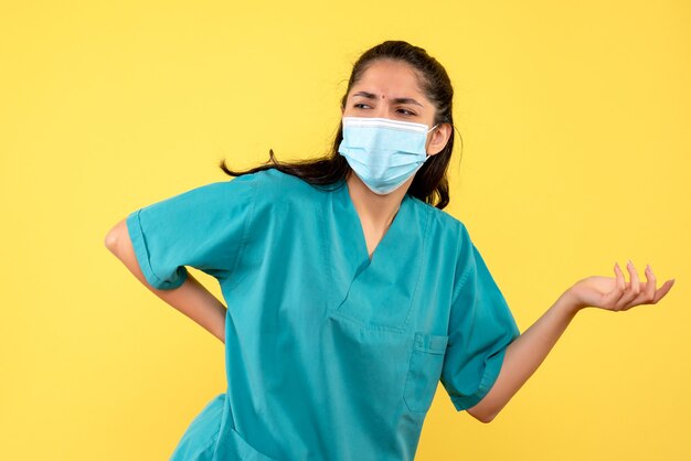 Vista frontal de la bonita doctora con máscara médica poniendo la mano en la cintura en la pared amarilla