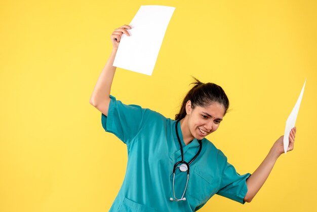 Vista frontal de la bonita doctora sosteniendo papeles con ambas manos en la pared amarilla
