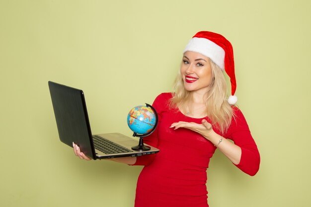 Vista frontal bonita mujer sosteniendo un pequeño globo terráqueo y usando la computadora portátil en la pared verde vacaciones emoción navidad nieve año nuevo colores