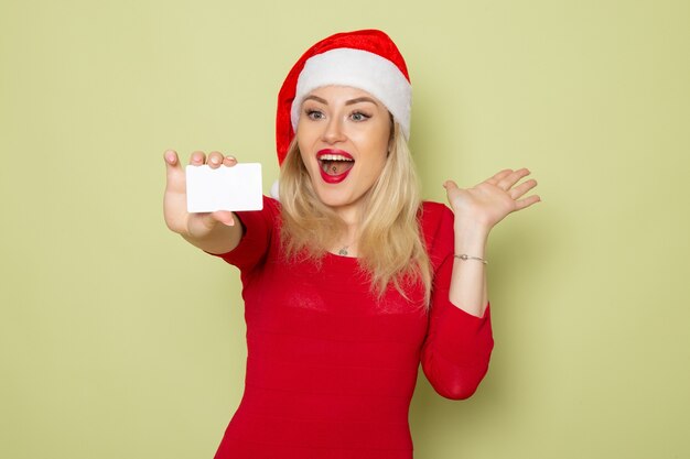 Vista frontal bonita mujer sosteniendo una tarjeta bancaria en la pared verde color Navidad nieve año nuevo vacaciones emociones