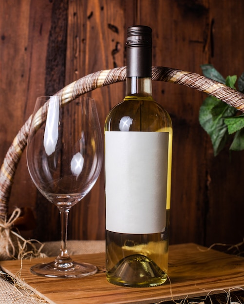 Una vista frontal de una botella de vino blanco junto con un vaso vacío en el mostrador de madera