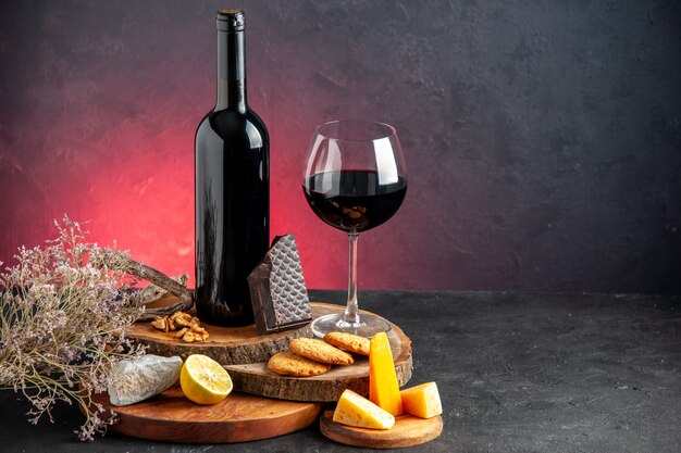 Vista frontal de la botella de vino negro vino tinto en vidrio queso cortado limón trozos de chocolate negro sobre tablas de madera rama de flores secas en la mesa roja lugar de copia