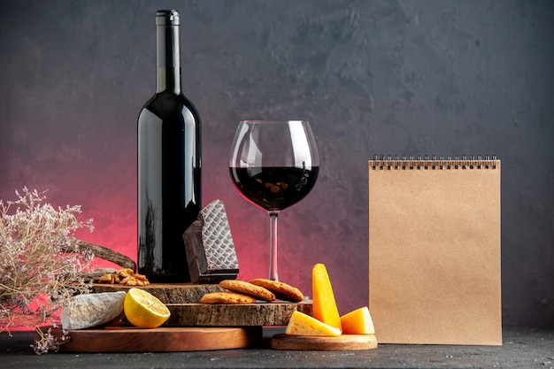 Vista frontal de la botella de vino negro vino tinto en vidrio queso cortado limón trozos de galletas de chocolate negro en tableros de madera cuaderno sobre mesa roja