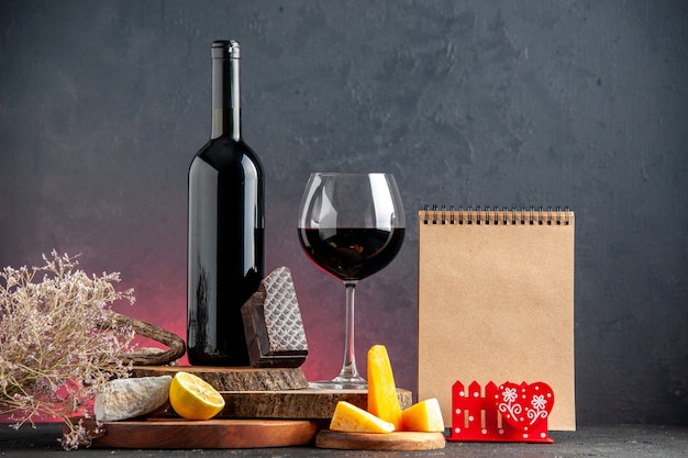 Vista frontal de la botella de vino negro vino en vidrio queso cortado limón un trozo de chocolate negro sobre tablas de madera Bloc de notas de rama de flor seca en la mesa roja