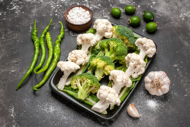 Vista frontal de brócoli crudo y coliflor en placa rectangular negra pimientos picantes verdes sal marina feykhoas ajo sobre fondo oscuro alimentos