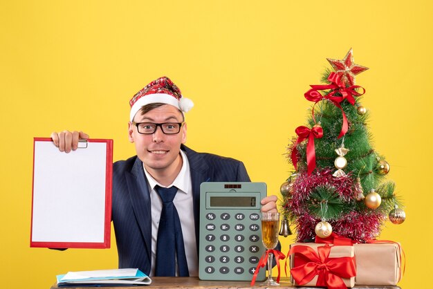 Vista frontal bsmiled hombre con gorro de Papá Noel sentado en la mesa cerca del árbol de Navidad y presenta sobre fondo amarillo