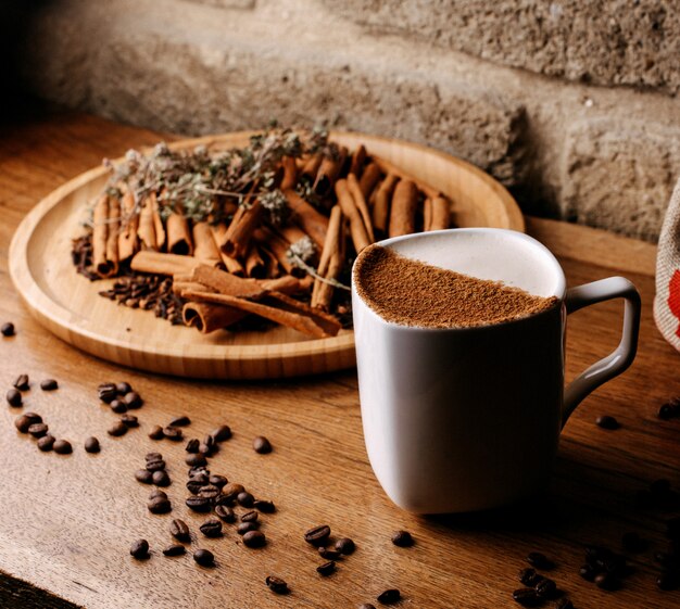 Vista frontal de café dentro de una taza blanca junto con semillas de café y canela