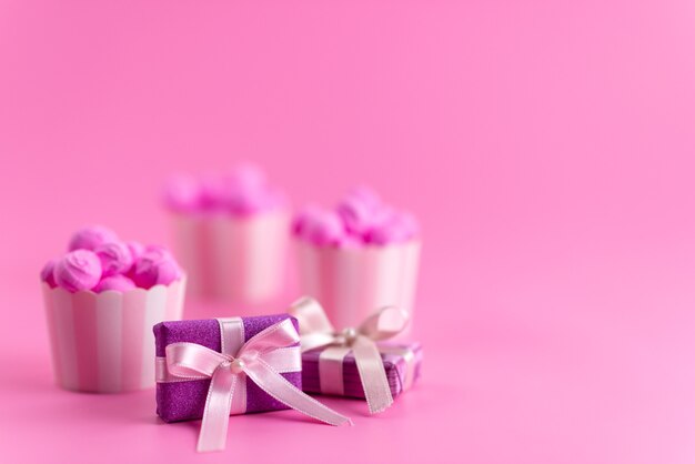 Una vista frontal de cajas de regalo de color púrpura junto con caramelos de color rosa en el escritorio rosa
