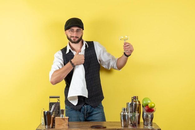 Vista frontal del camarero masculino frente a la mesa con agitadores limpiando vasos en la pared amarilla bar club nocturno de bebidas alcohólicas para jóvenes