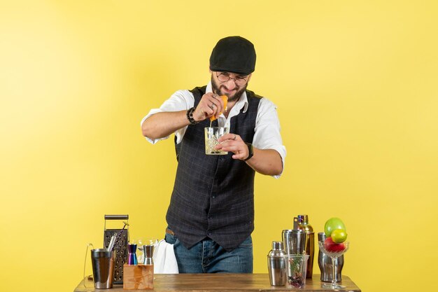 Vista frontal del camarero masculino frente a la mesa con agitadores preparando bebida exprimiendo naranja en la pared amarilla bar club nocturno de bebidas alcohólicas juveniles