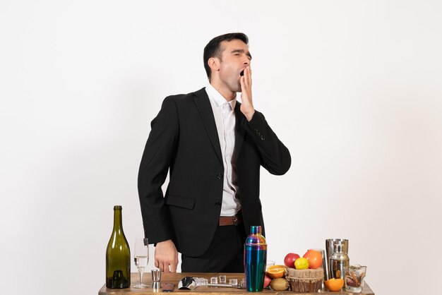 Vista frontal del camarero masculino de pie delante de la mesa con bebidas bostezando en la pared blanca noche barra de alcohol del club de bebidas masculinas