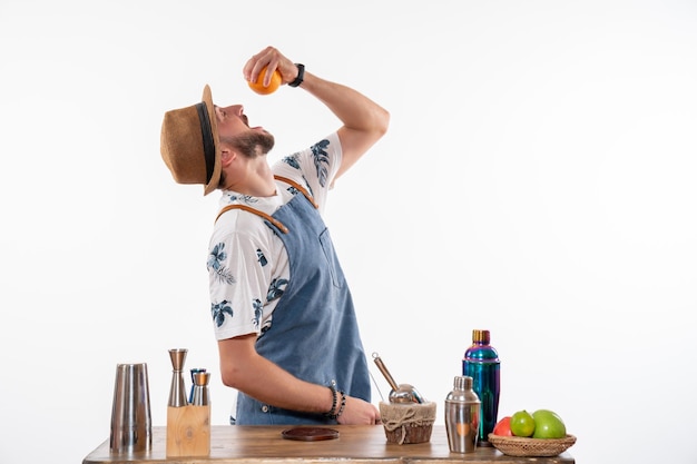 Vista frontal del camarero masculino probando naranja y haciendo bebidas en la pared blanca barra de trabajo club de alcohol servicio de bebidas nocturnas