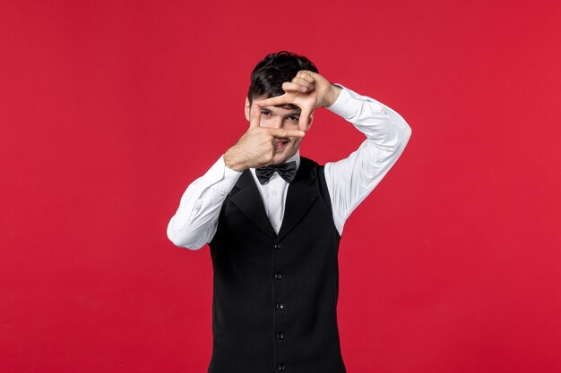 Vista frontal del camarero masculino sonriente en un uniforme con mariposa en el cuello haciendo gesto ok sobre fondo rojo.