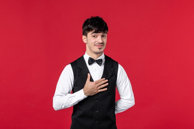 Vista frontal del camarero masculino sonriente en uniforme con pajarita en el cuello y agradeciendo a alguien en la pared roja