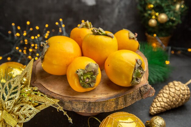 Vista frontal de los caquis frescos alrededor de los juguetes de navidad sobre fondo oscuro, jugo exótico exótico de frutas tropicales.