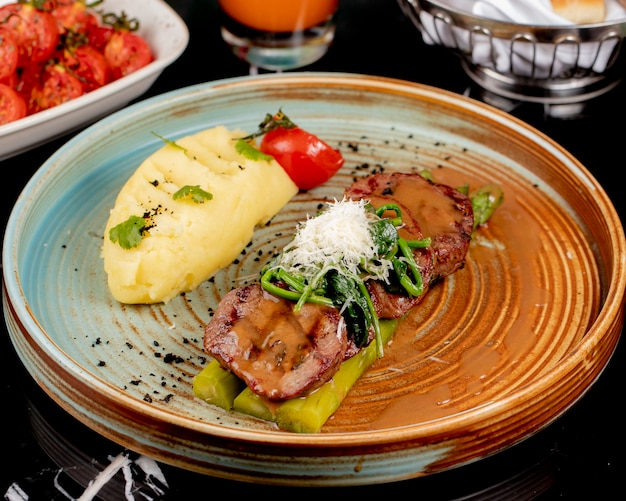 Vista frontal de carne frita sobre espárragos con puré de papas y hierbas en un plato