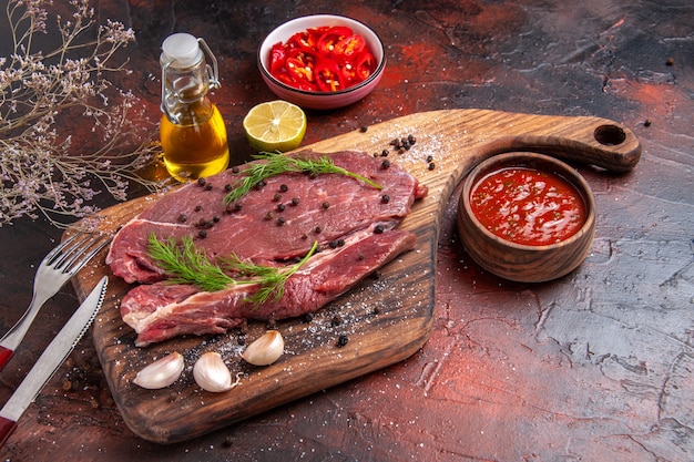 Vista frontal de la carne roja en la tabla de cortar de madera y el ajo pimiento verde tenedor y cuchillo sobre fondo oscuro