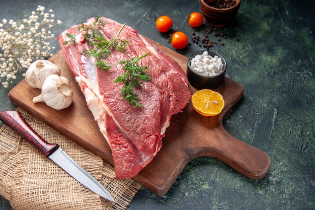 Vista frontal de carnes rojas crudas frescas, ajos verdes, sal de limón en un cuchillo de tabla de cortar de madera marrón en una toalla de color nude, tomates, pimienta sobre fondo de color oscuro